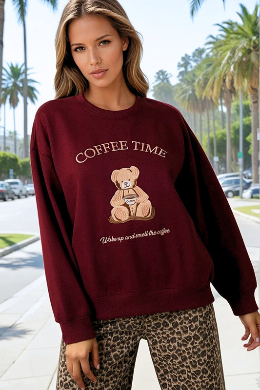 Woman wearing a maroon sweatshirt with a teddy bear graphic and 'Coffee Time' text, standing on a street with palm trees in the background.