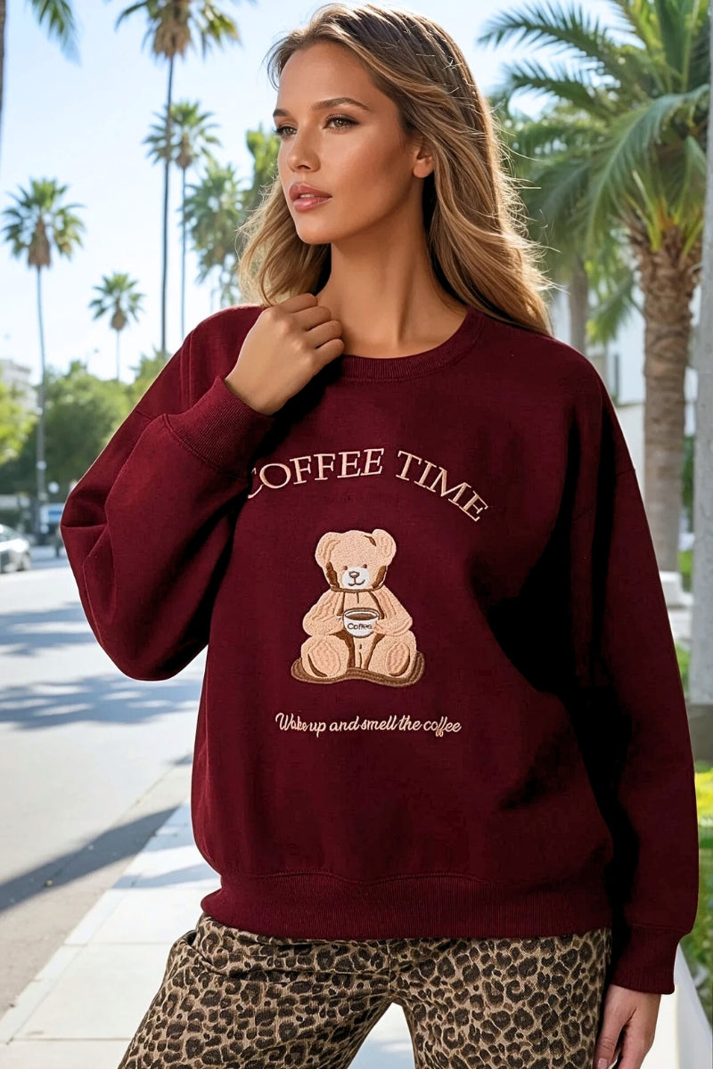 Woman wearing a maroon sweatshirt with a bear graphic and 'Coffee Time' text on a street with palm trees.
