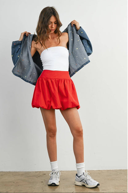 Woman wearing a white top, red skirt, and gray jacket against a plain background