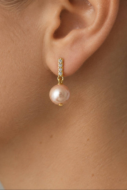 A close-up of a model's ear wearing a gold huggie hoop earring with a vertical row of small clear stones and a dangling pink pearl.