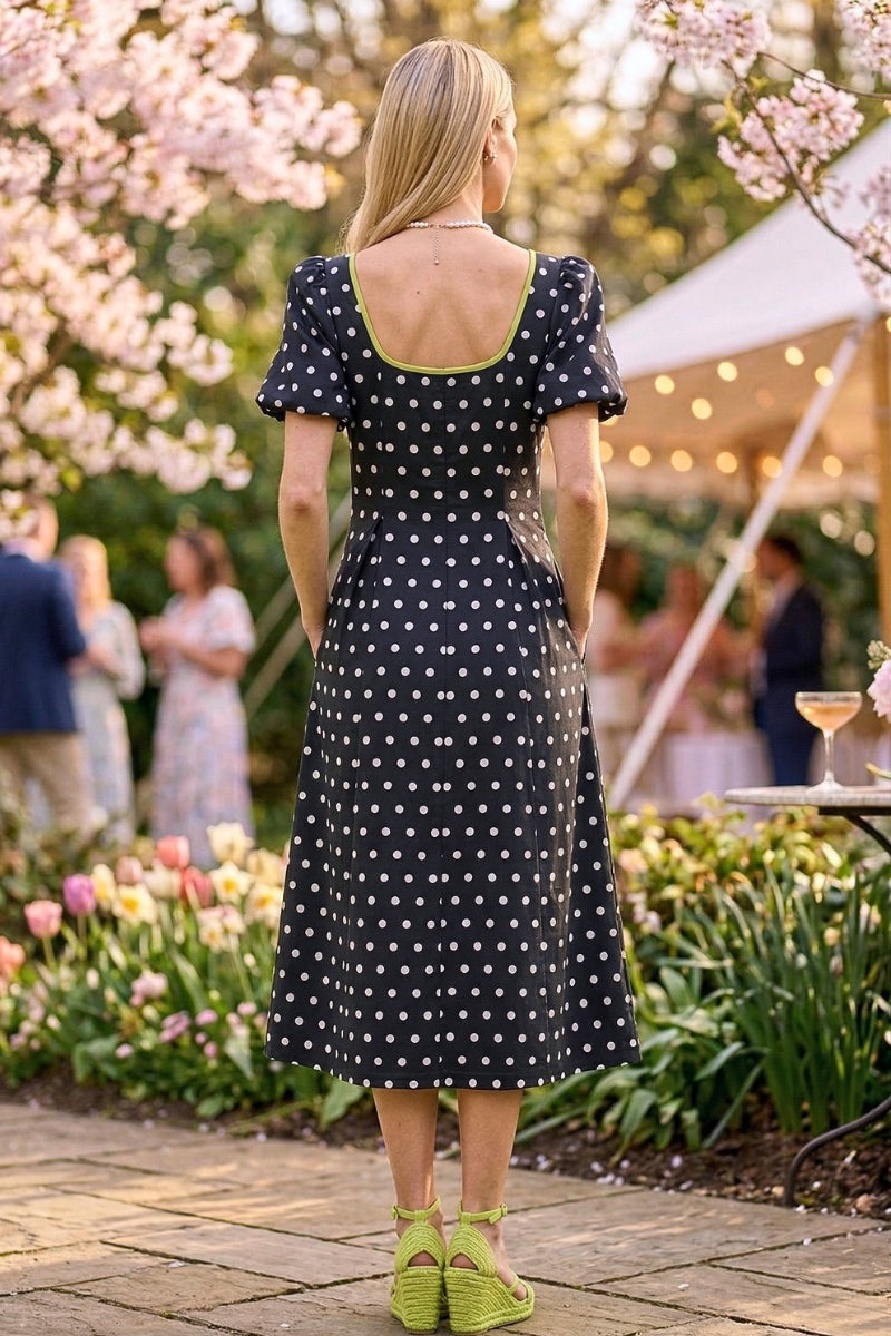 Woman in a black polka dot dress standing in a garden with cherry blossoms and people in the background.