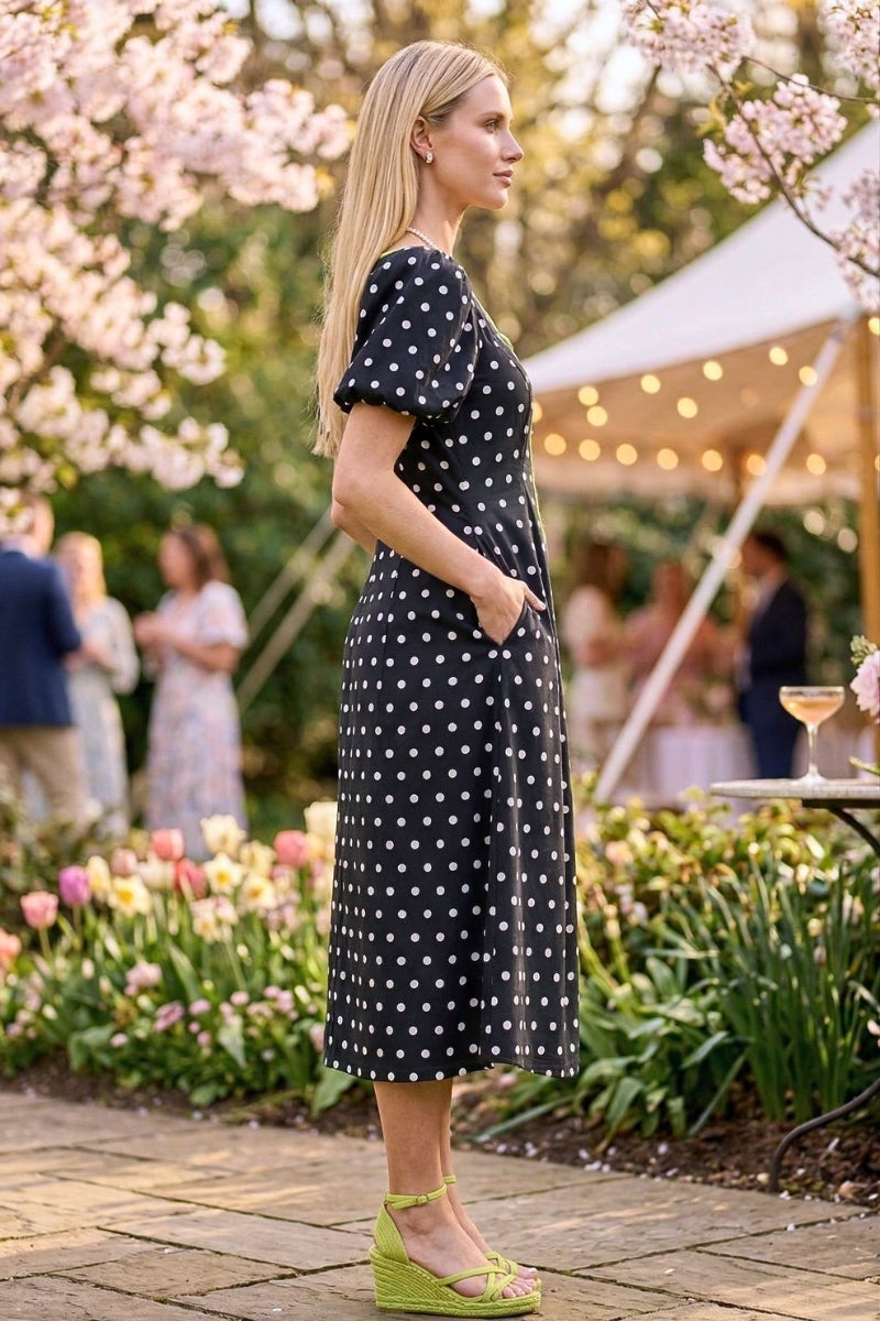 Woman in a black polka dot dress standing outdoors with cherry blossoms and a garden in the background.