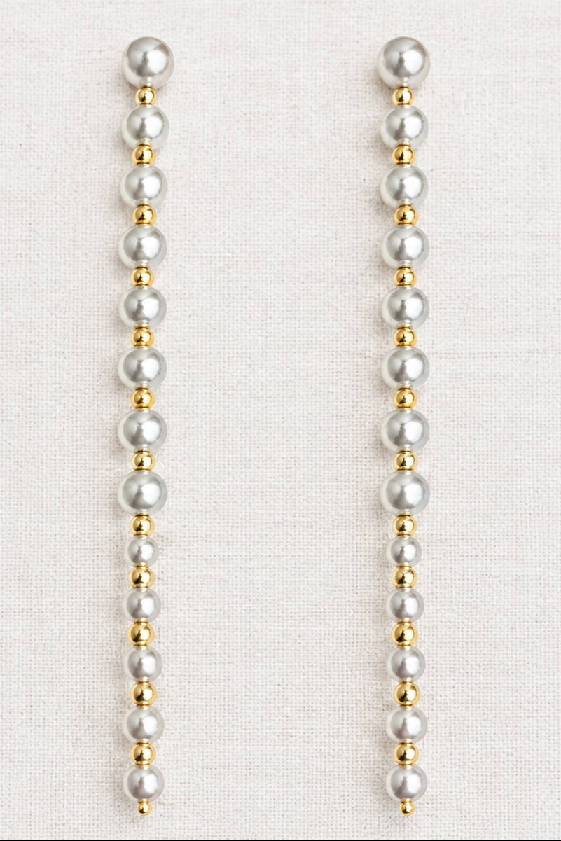 A pair of long drop earrings made of white faux pearls and small gold spacer beads, displayed on a textured off-white fabric background.