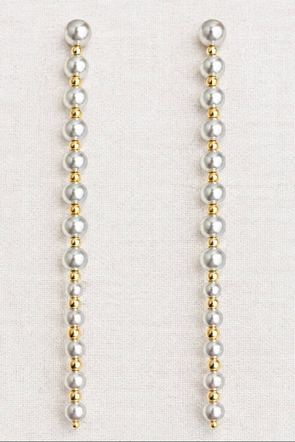 A pair of long drop earrings made of white faux pearls and small gold spacer beads, displayed on a textured off-white fabric background.