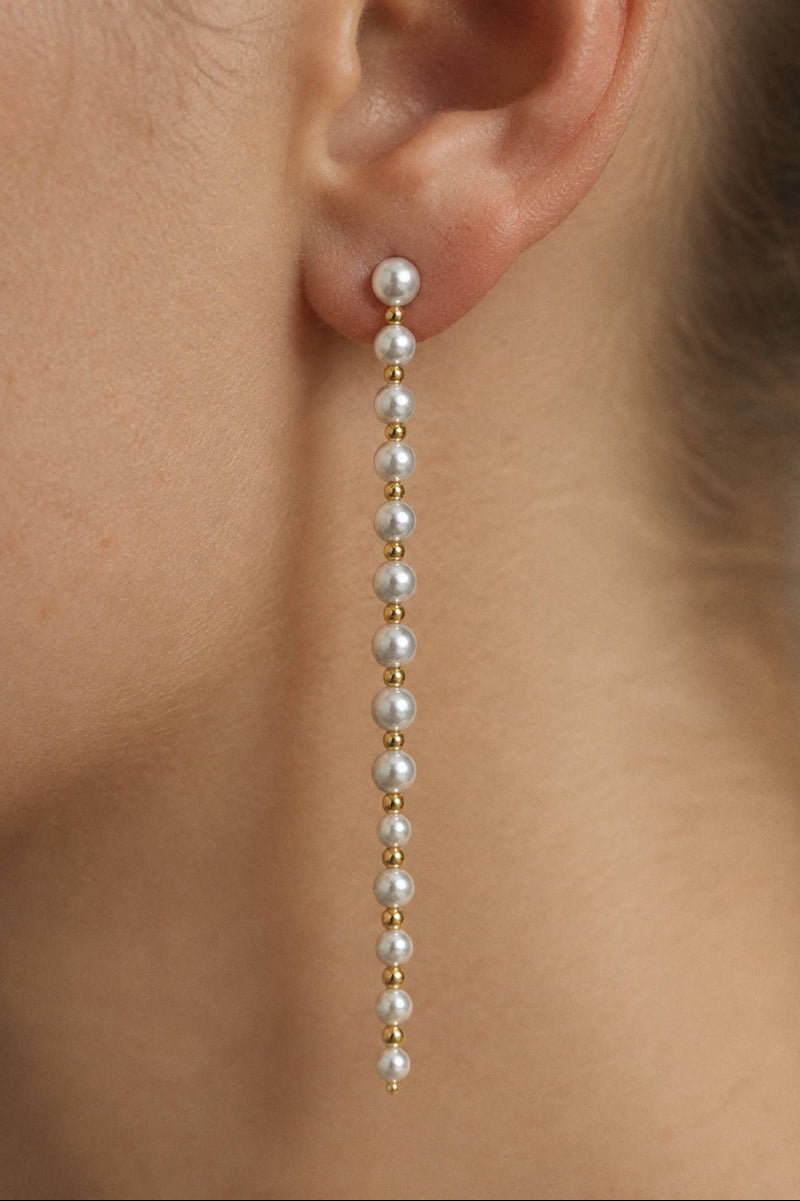 A close-up of a model's ear wearing a long drop earring made of small white pearls and gold spacer beads.