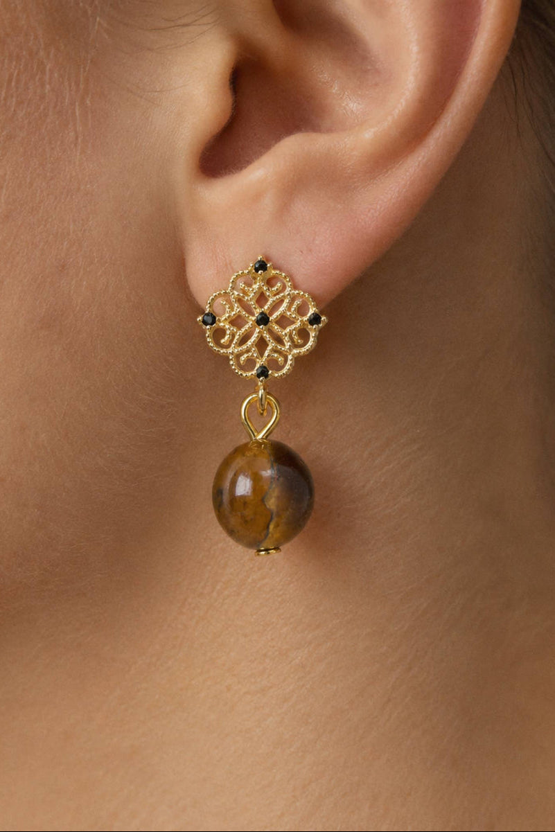 A close-up of a gold dangle earring worn on a person's ear. The earring features an intricate filigree top with tiny dark blue stones and a smooth, polished brown tiger's eye bead dangling below.
