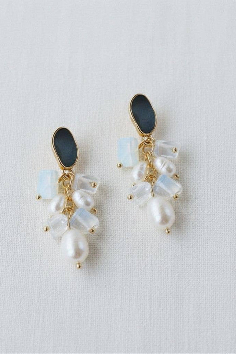 A pair of gold-tone dangle earrings featuring a dark gray oval stone, white baroque pearls, and milky white cubic zirconia accents, displayed on a textured white fabric background.