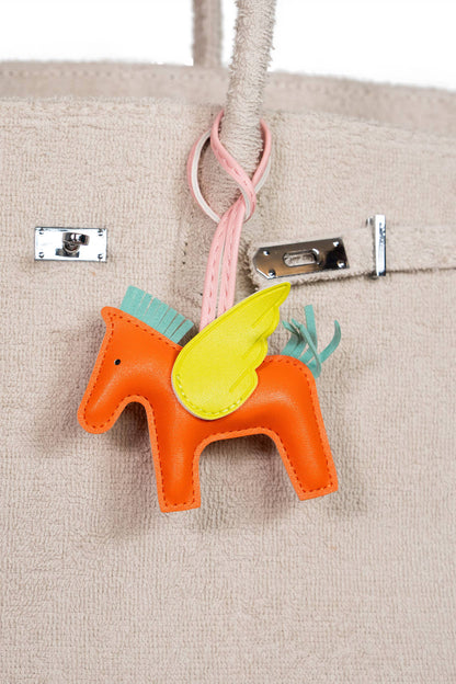 Orange horse-shaped keychain with yellow wings attached to a beige handbag.