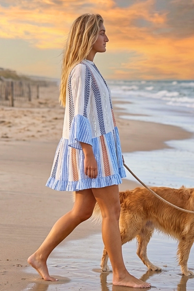 Woman in a blue and white striped dress walking a dog on a beach at sunset.
