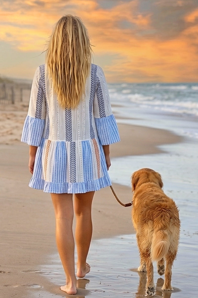 Woman in a blue and white striped dress walking a dog on a beach at sunset.