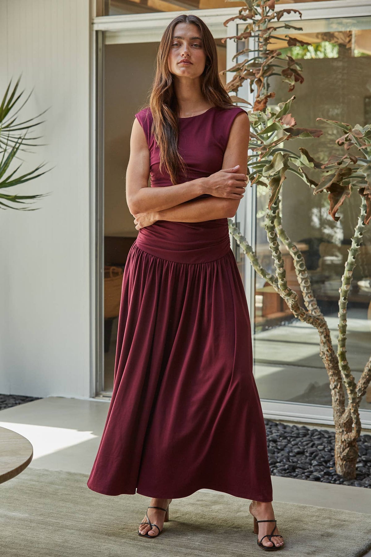 A woman modeling a sleeveless, high-waisted maxi dress in a deep burgundy or maroon color, standing in a modern patio setting with plants and glass doors.