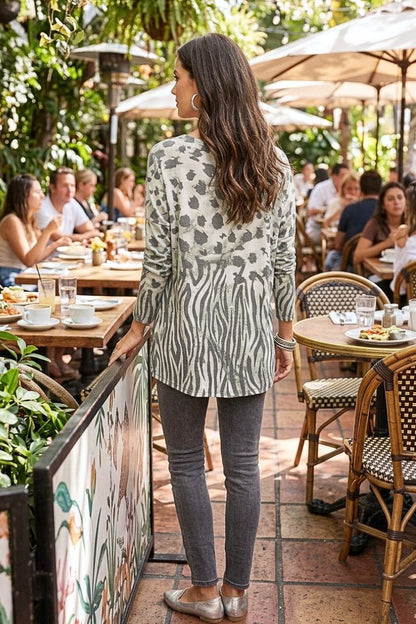 A woman stands at an outdoor patio table wearing a long-sleeve zebra-print tunic top with gray skinny jeans and silver flats, photographed in a real cafe environment with seated people and umbrellas in the background.