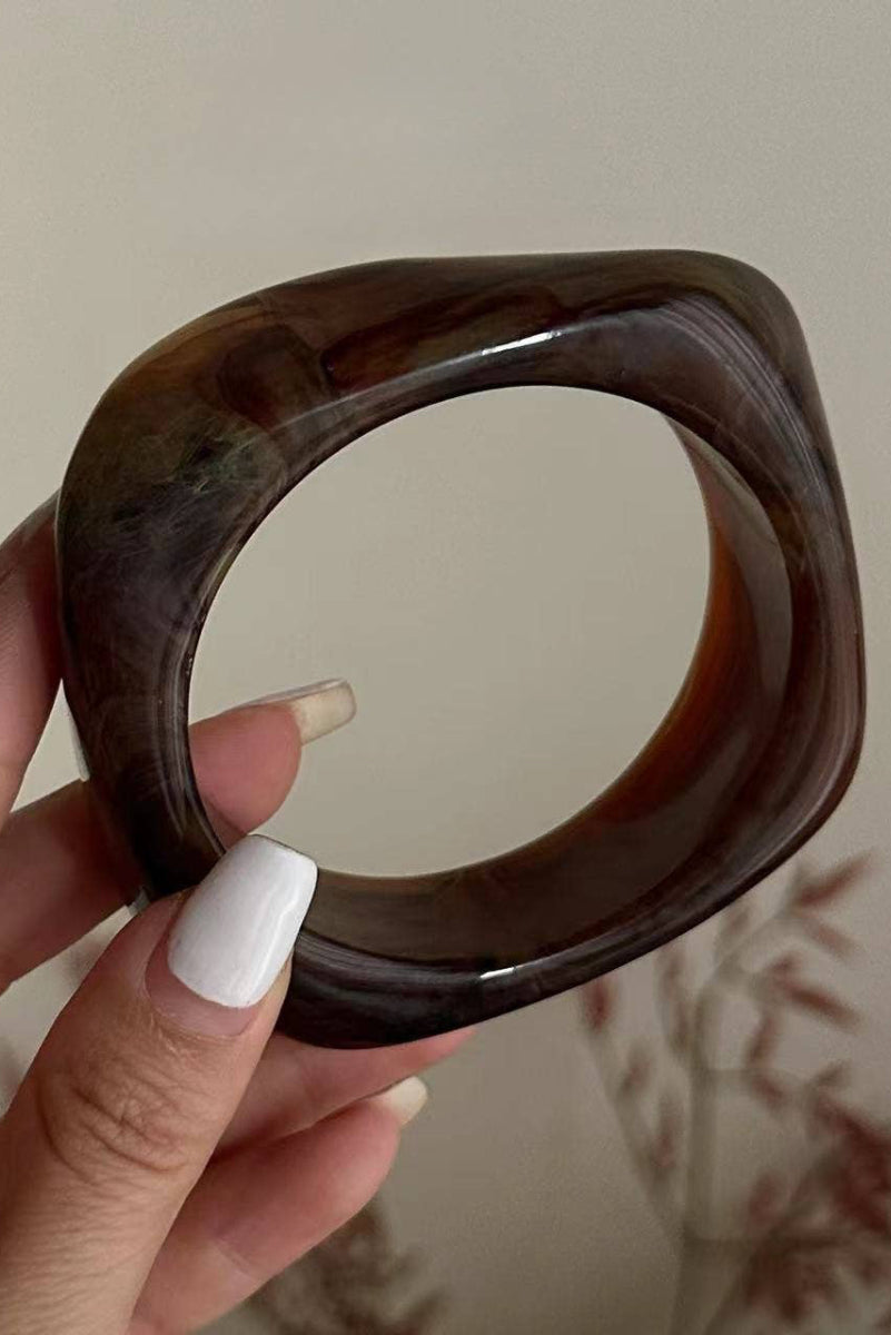 Brown bangle held by a hand with white nail polish against a neutral background