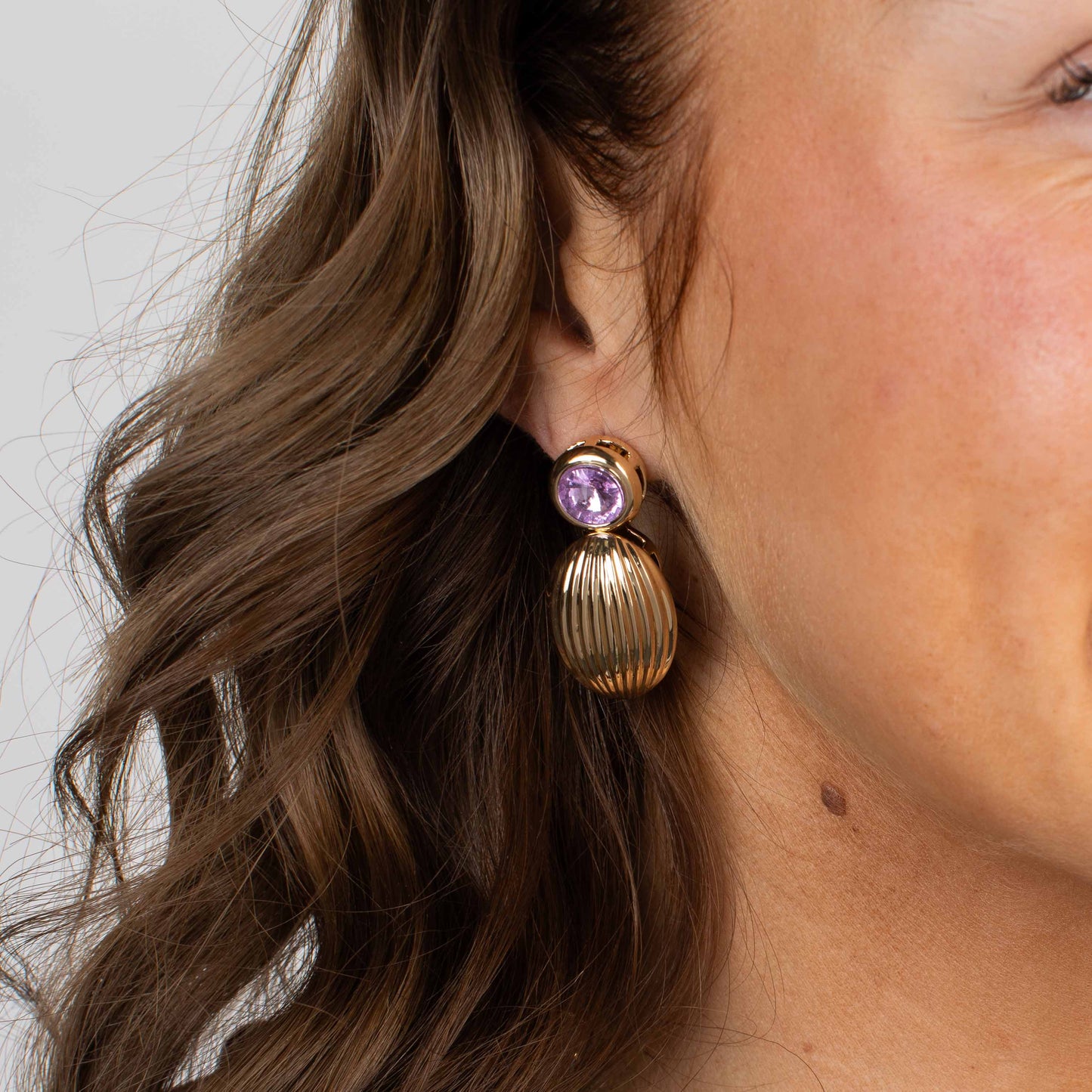Gold earring with a purple gemstone worn by a person with wavy hair.