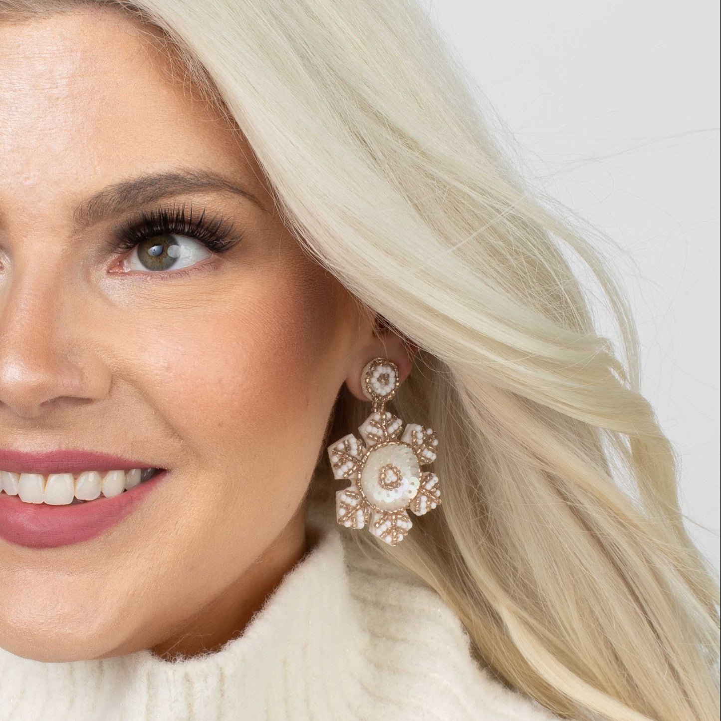 Woman with blonde hair wearing a white turtleneck and floral earrings against a light background