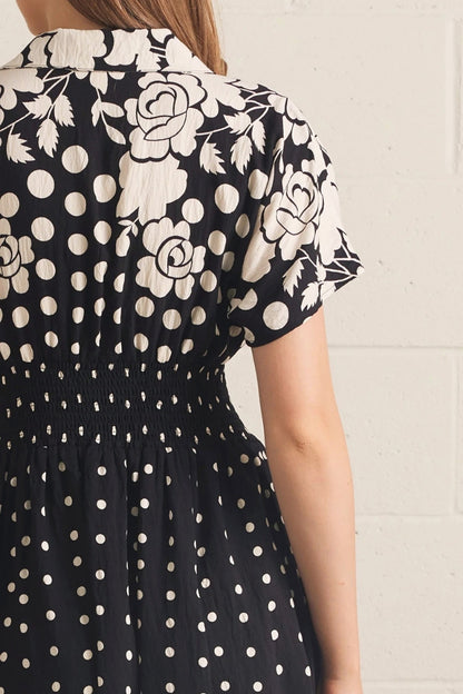 A close-up back view of a woman wearing a black dress with a white floral rose print on the upper back and shoulders, and a white polka dot pattern on the skirt, against a light brick wall.