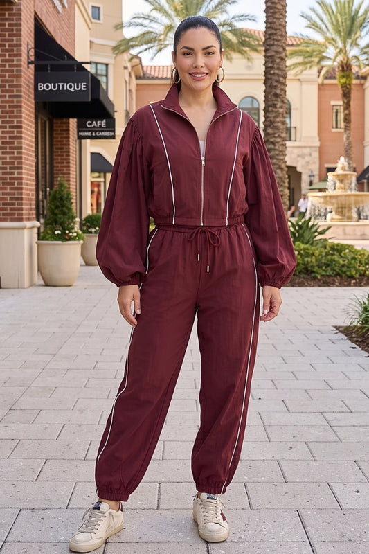 A woman stands outdoors on a paved walkway wearing a matching burgundy zip-up jacket and jogger pants set with thin white side stripes. The setting includes storefronts, palm trees, and a fountain in the background.