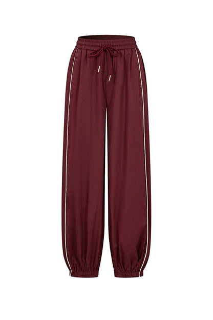 A pair of relaxed-fit jogger pants in a deep burgundy color, featuring white stripes down the sides and a matching drawstring waistband, shown against a plain white background.