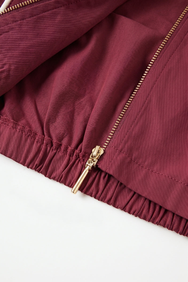 Close-up of a burgundy or deep wine-colored jacket or fabric panel with a gold-tone zipper and elasticized hem, laid on a white background.