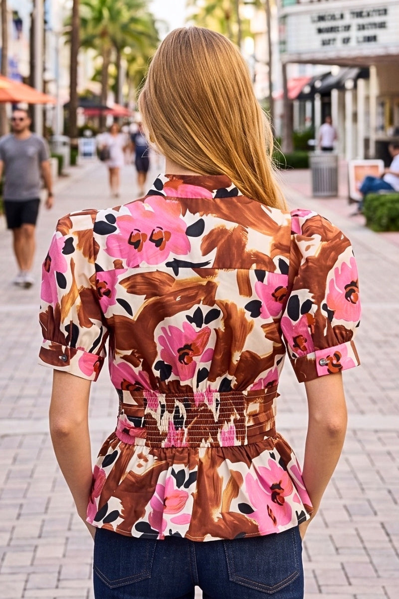 A woman viewed from behind wearing a short-sleeve peplum blouse with a brown, pink, and black floral pattern, standing on a paved outdoor walkway.