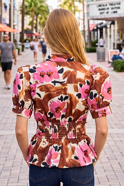 A woman viewed from behind wearing a short-sleeve peplum blouse with a brown, pink, and black floral pattern, standing on a paved outdoor walkway.