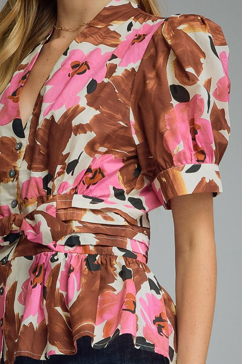 A close-up of a woman wearing a short-sleeve wrap-style blouse with a brown, pink, and cream floral print. The blouse features puff sleeves, a wrap front with visible buttons, and a tied waist belt.