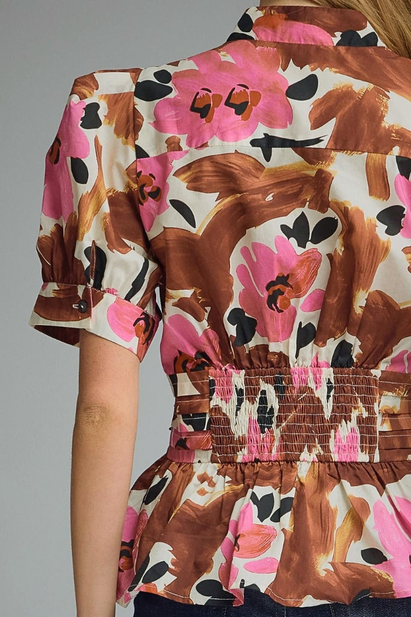 A close-up of a woman wearing a short-sleeve blouse with a brown, pink, and black abstract floral print on a cream background. The blouse features a smocked waistband and a peplum hem.