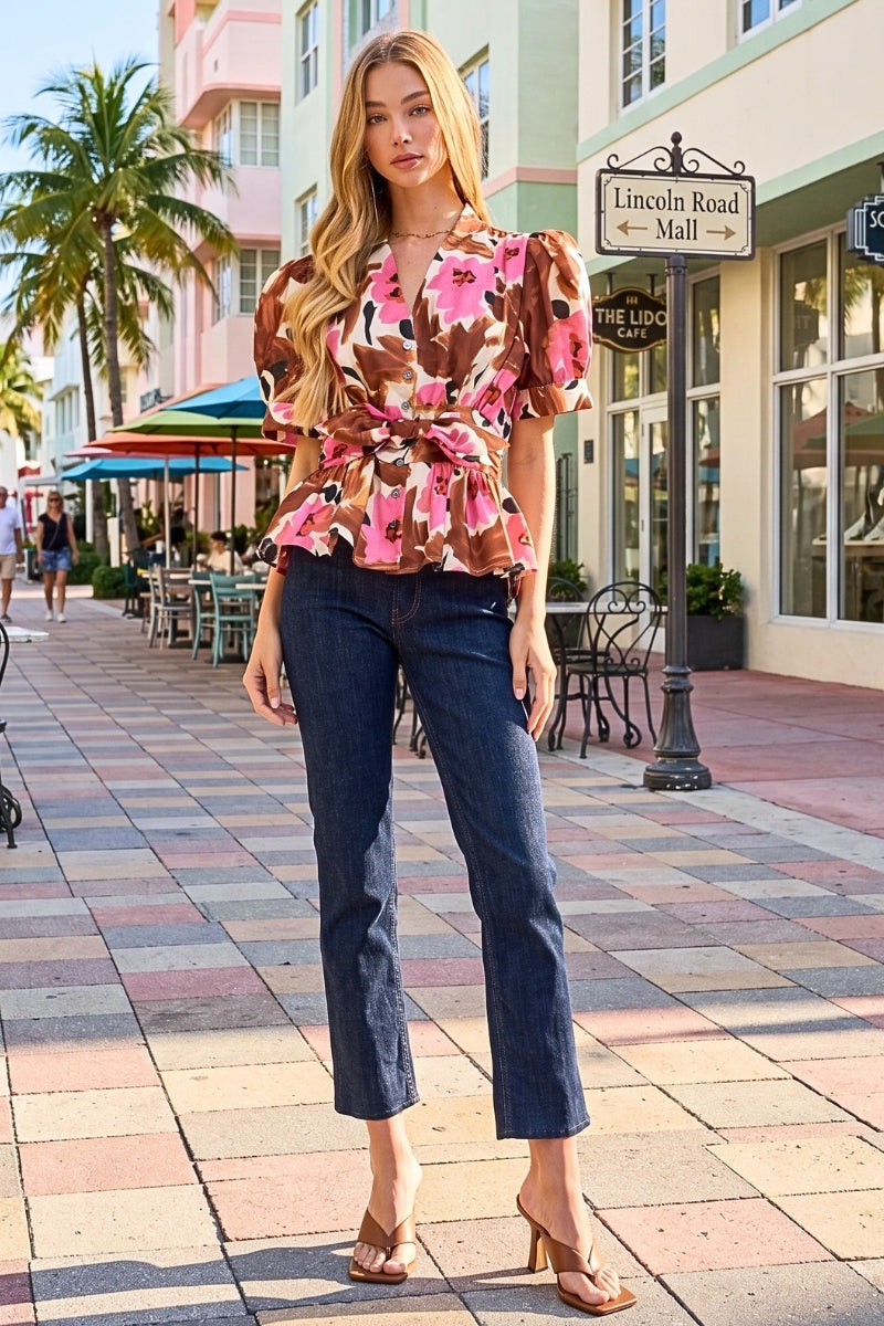 A woman stands on a sunlit pedestrian street wearing a brown floral puff-sleeve blouse with a tie waist and dark blue cropped jeans, paired with tan heeled sandals. Buildings, palm trees, and storefronts are visible in the background.