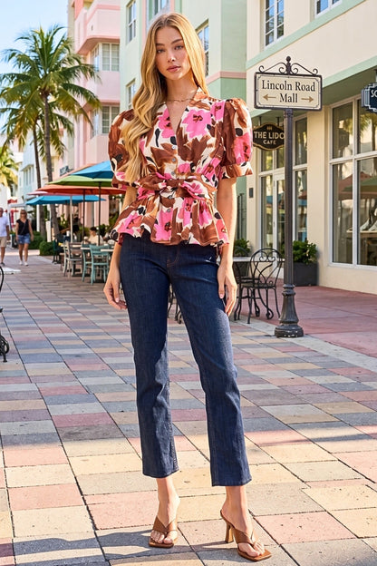 A woman stands on a sunlit pedestrian street wearing a brown floral puff-sleeve blouse with a tie waist and dark blue cropped jeans, paired with tan heeled sandals. Buildings, palm trees, and storefronts are visible in the background.