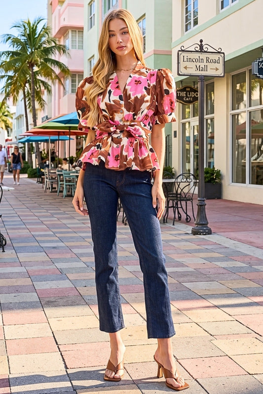 A woman stands on a sunlit pedestrian street wearing a brown floral puff-sleeve blouse with a tie waist and dark blue cropped jeans, paired with tan heeled sandals. Buildings, palm trees, and storefronts are visible in the background.