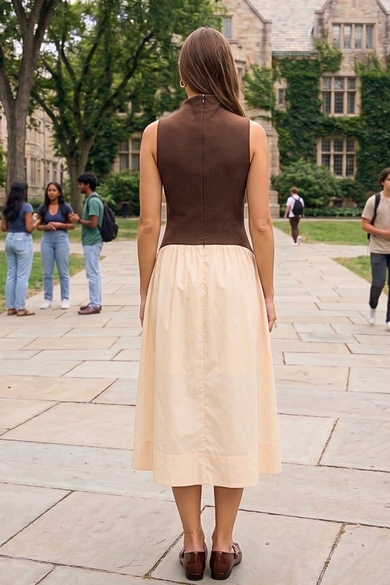 A woman viewed from behind wearing a sleeveless two-tone dress with a dark brown bodice and a light beige skirt, standing on a stone walkway in front of a college building with trees and other people in the background.