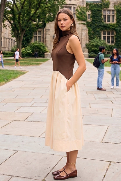 A woman stands on a stone walkway in front of a large ivy-covered stone building, wearing a sleeveless midi dress with a brown bodice and a light beige skirt, paired with brown flat shoes.