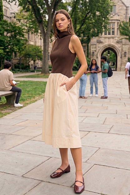 A woman stands on a stone walkway in front of a university-style building, wearing a sleeveless brown mock-neck top and a light cream midi skirt with side pockets, paired with dark brown flats.