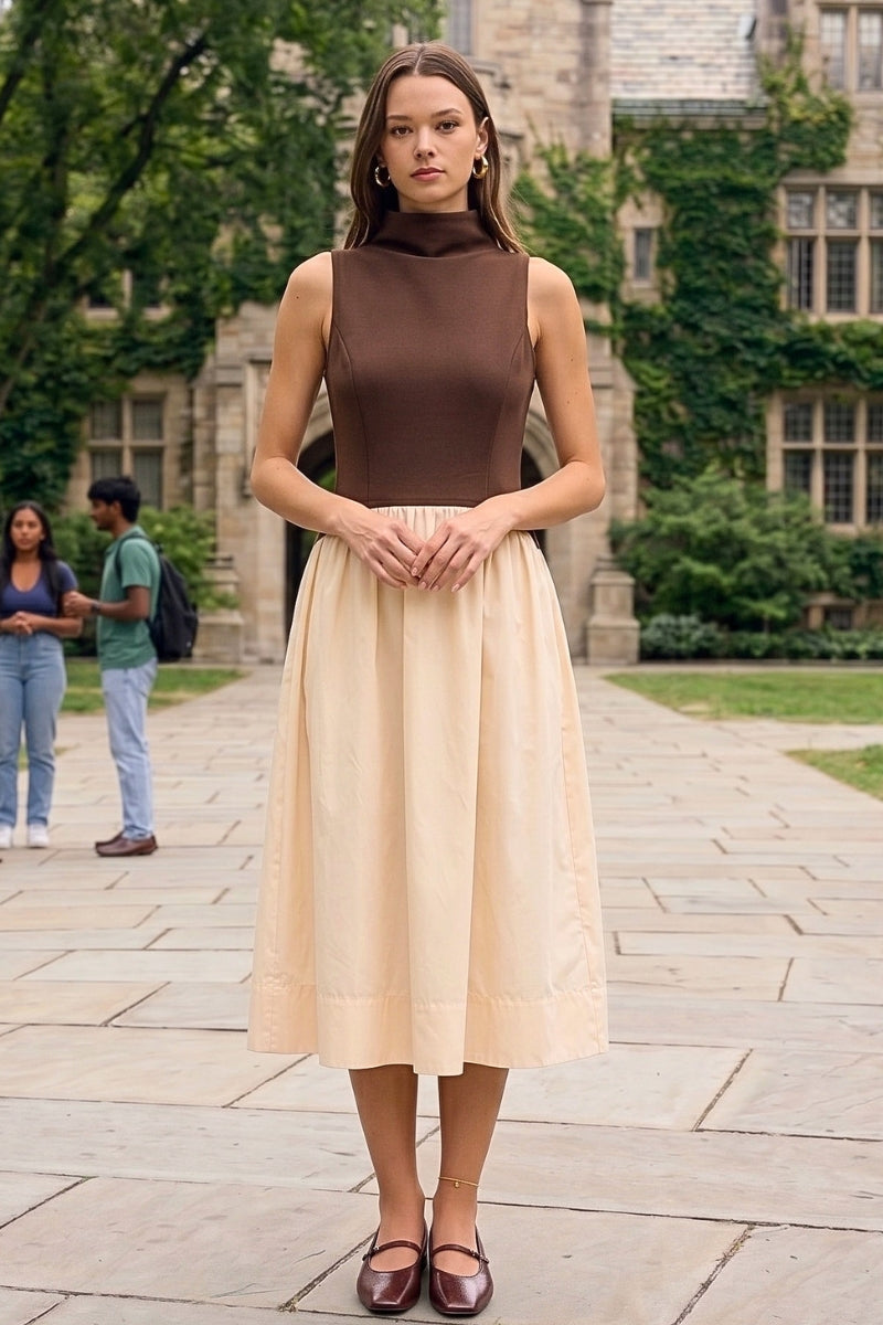 A woman stands on a stone walkway in front of a historic stone building with ivy, wearing a sleeveless mock-neck dress with a dark brown bodice and a light beige skirt, paired with dark brown flats.