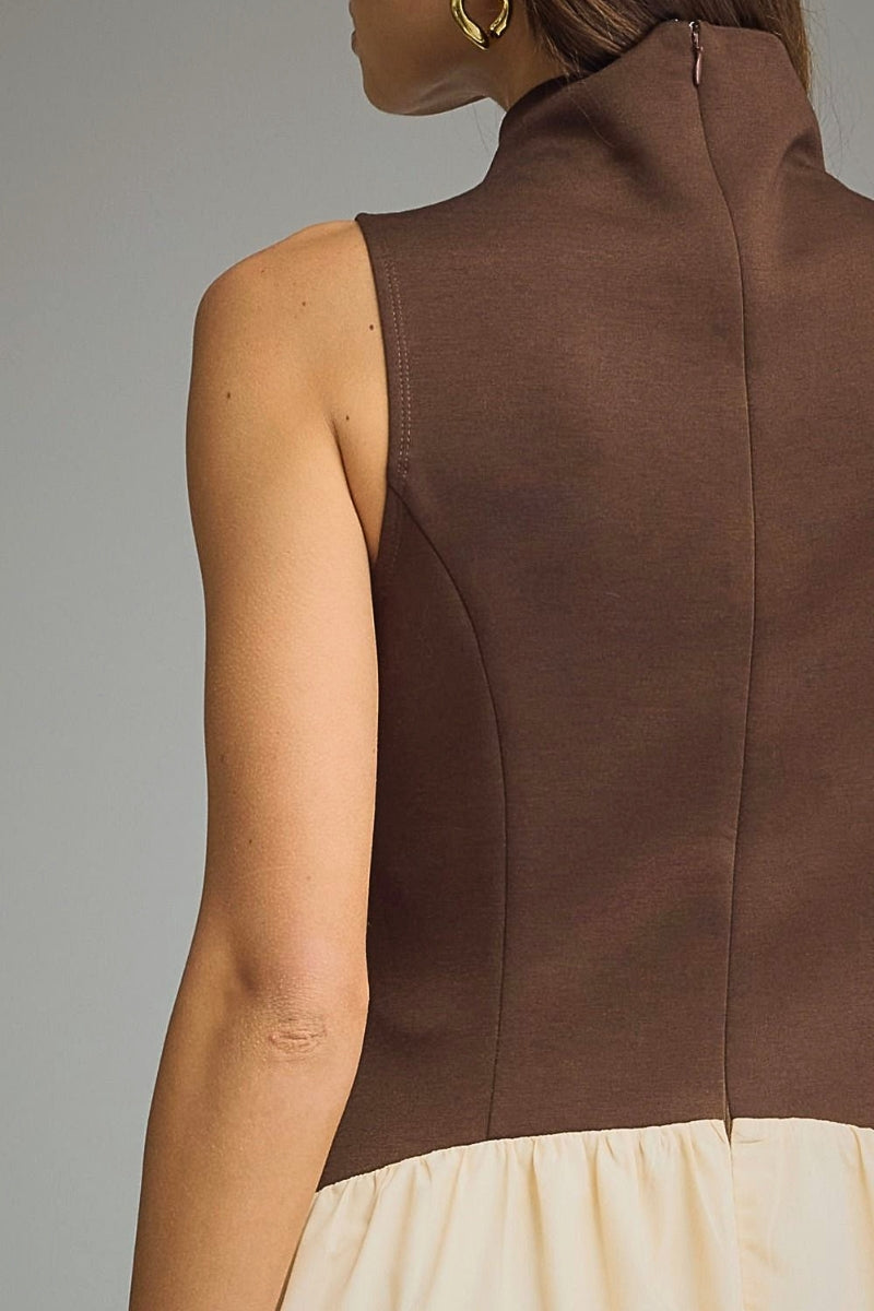 Close-up back view of a model wearing a sleeveless high-neck dress with a dark brown upper section and a cream-colored lower ruffled section, set against a plain gray studio background.