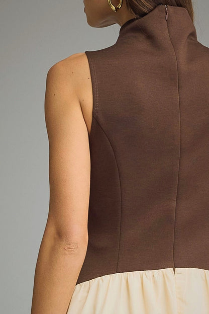 Close-up back view of a model wearing a sleeveless high-neck dress with a dark brown upper section and a cream-colored lower ruffled section, set against a plain gray studio background.