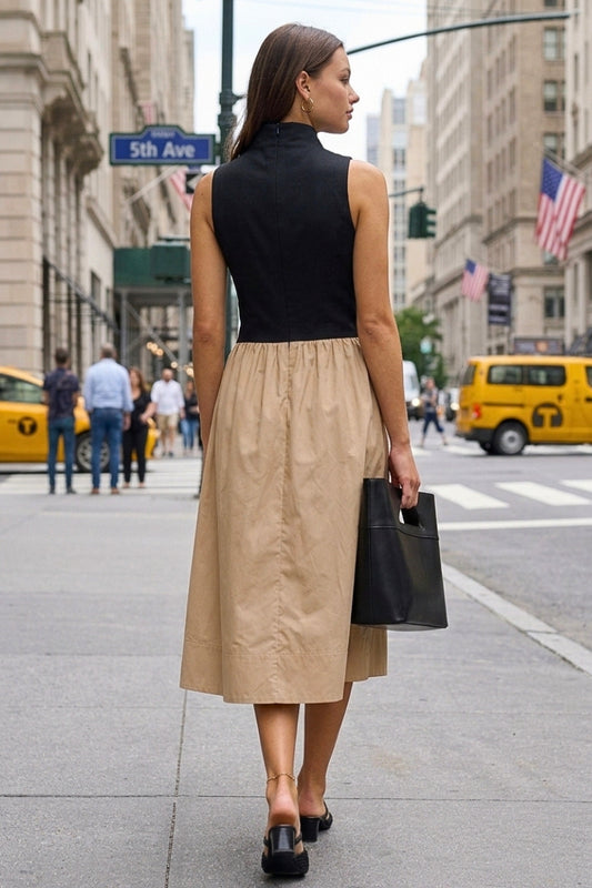 A woman walking on a city street viewed from behind, wearing a sleeveless black mock-neck top and a knee-length beige skirt, carrying a black handbag.