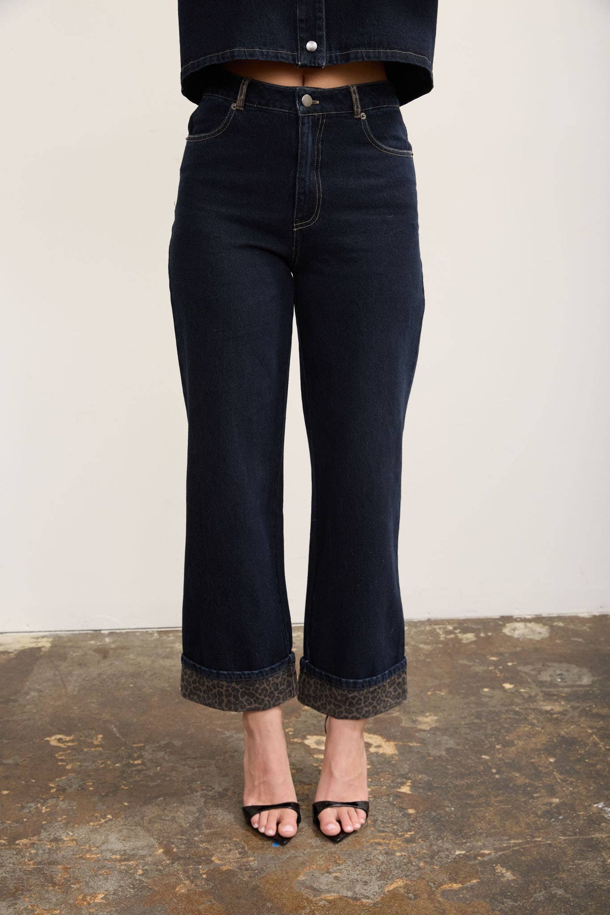 A person wearing dark indigo cropped flare jeans with rolled cuffs and a textured hem, paired with black heeled sandals against a plain light wall and rustic floor.