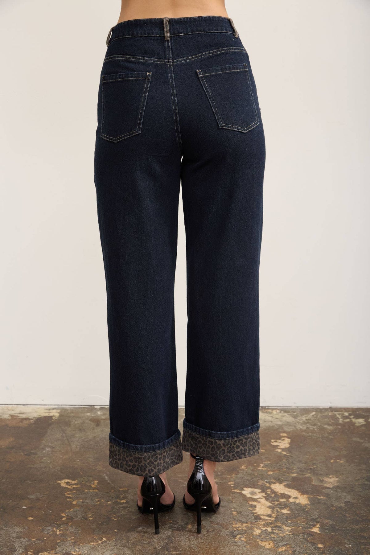 Back view of a person wearing dark blue wide-leg cropped jeans with rolled cuffs and black high heels, standing against a plain light wall and textured floor.