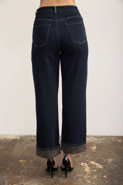 Back view of a person wearing dark blue wide-leg cropped jeans with rolled cuffs and black high heels, standing against a plain light wall and textured floor.