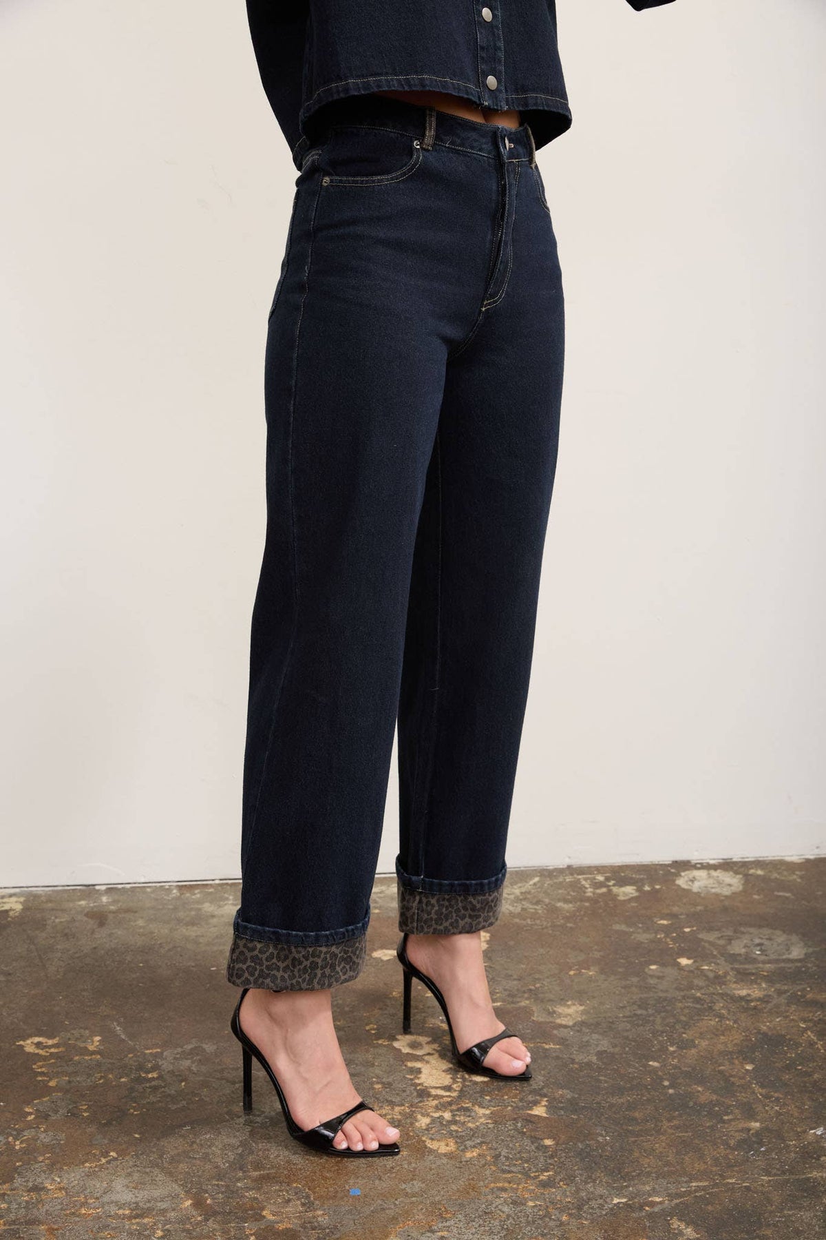 A person wearing dark indigo cropped jeans with rolled cuffs and black high-heeled sandals, standing against a plain light wall on a textured concrete floor.