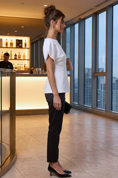 A woman stands in profile in a modern high-rise office lobby, wearing a white short-sleeve peplum-style top and black trousers with black heels.