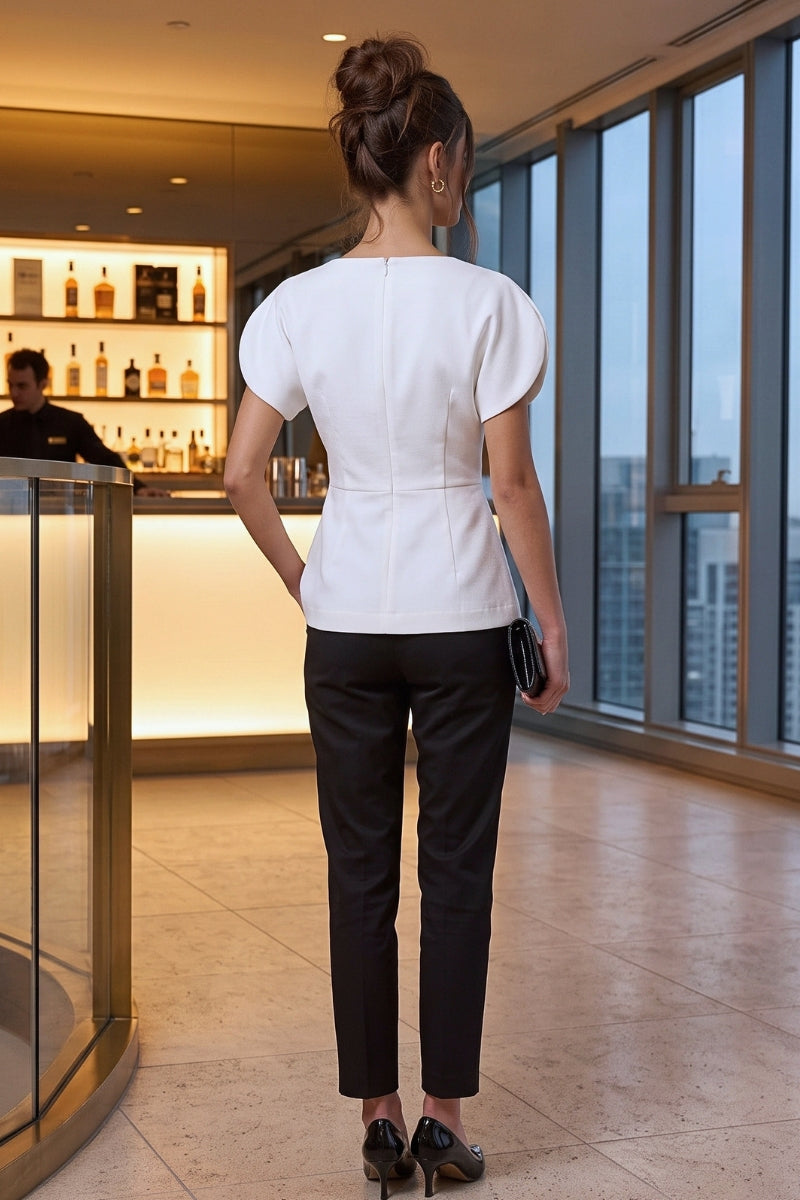 A woman viewed from behind wearing a white short-sleeve peplum-style top and black trousers, standing in a modern indoor setting with large windows and a bar visible in the background.
