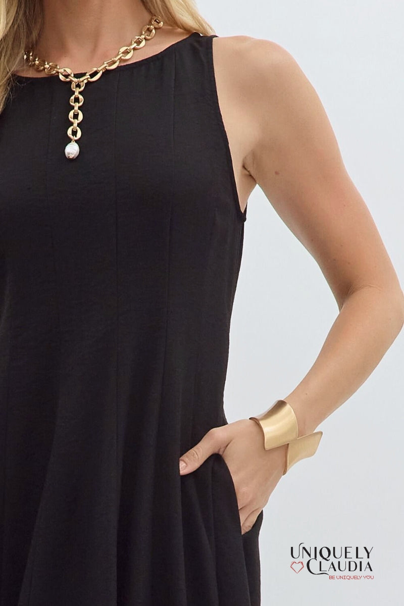 Woman wearing a black sleeveless dress with a gold necklace and bracelet on a light gray background.