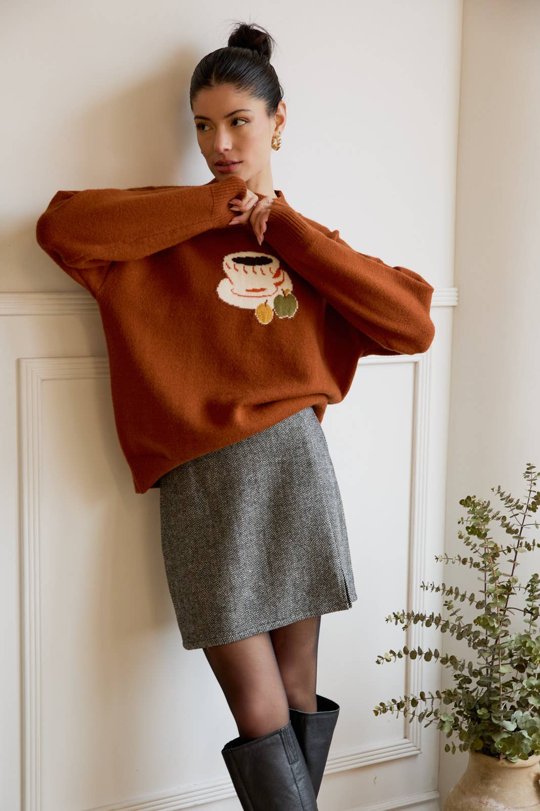 Woman wearing a brown sweater with a coffee design, gray skirt, and black boots against a white wall.