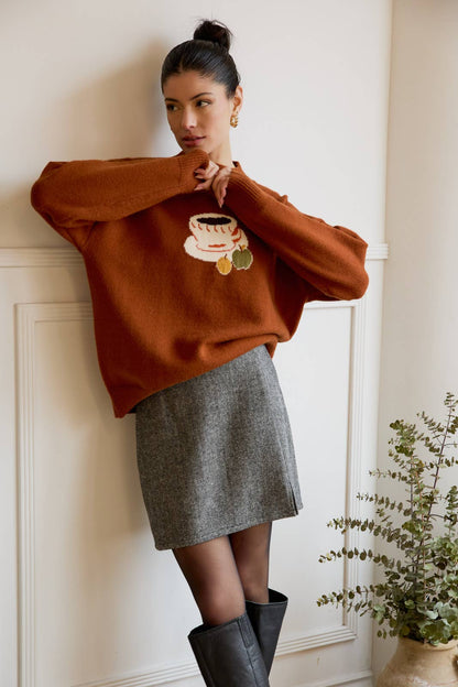 Woman wearing a brown sweater with a coffee design, gray skirt, and black boots against a white wall.