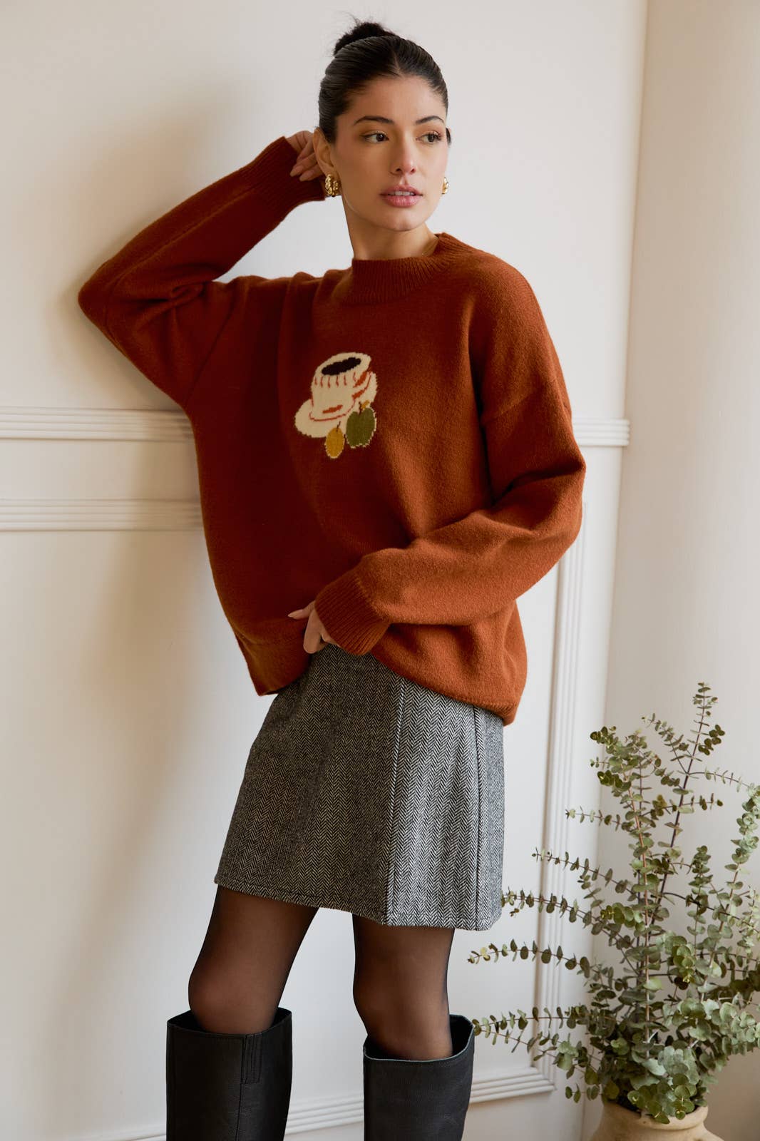 Woman wearing a brown sweater with a coffee design, gray skirt, and black boots indoors.