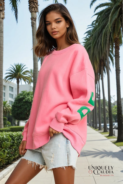 Woman wearing a pink sweatshirt with green text in an outdoor setting with palm trees.