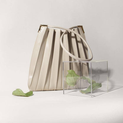 Beige pleated handbag and clear cube with green leaves on a light gray background