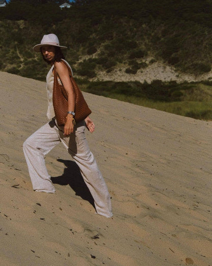 Woman walking on a sandy dune wearing a hat, beige outfit, and saddle bag.
